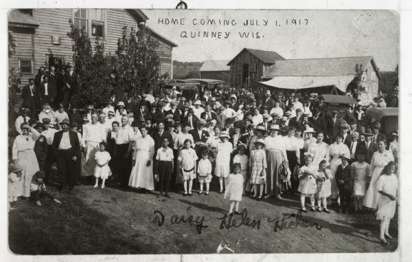 View of a large group of people gathered for a Homecoming celebration. A young woman in the foreground is identified as Daisy Helen Hicken.