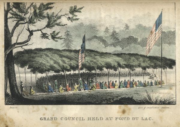 View of large gathering of people among trees. A crowd is gathered in a semi-circle facing a table where two people are standing. Two U.S flags fly over the proceedings.