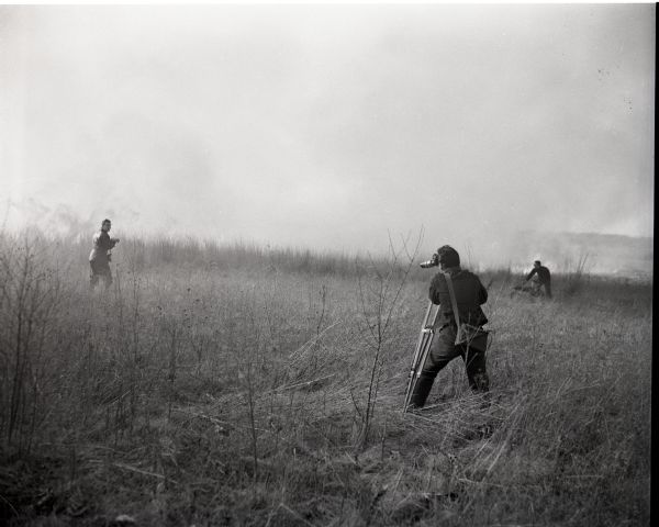 Disney film crew shooting a prescribed burn at the University of Wisconsin-Madison Arboretum for inclusion in the film <i>The Vanishing Prairie</i>. A cameraman with a camera on a tripod films in the foreground while other crew members work closer to the advancing smoke and fire.