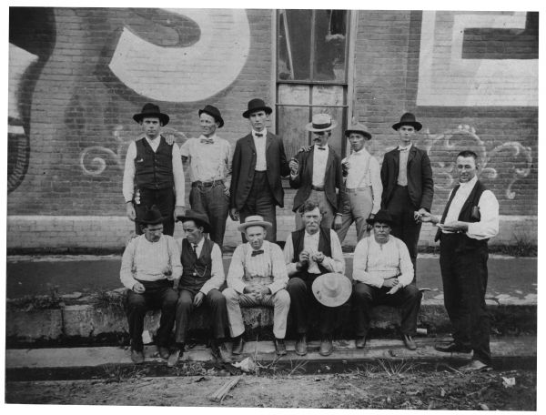 Probable group of pearl buyers posed curbside. The partial wall of the building behind the men has a window and the bricks are painted with an advertisement.