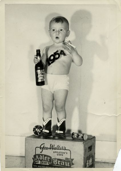 A young Jimmy Clark dressed as Baby New Year for 1954. He stands on top of a case of Adler Bräu wearing cowboy boots, underwear, and a sash reading 1954. He also holds a cigarette and a bottle of Adler Bräu. Two toys lay on the box next to his boots.