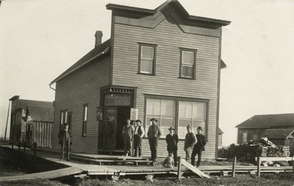 Eight men standing outside a corner tavern. The men are standing on a plank sidewalk with a dog. The is a Blatz Beer sign hanging outside the door.