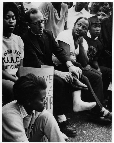 Father James Groppi sitting with several of his fellow marchers, some in Milwaukee N.A.A.C.P. Youth Council shirts. Groppi has taken off one of his shoes.