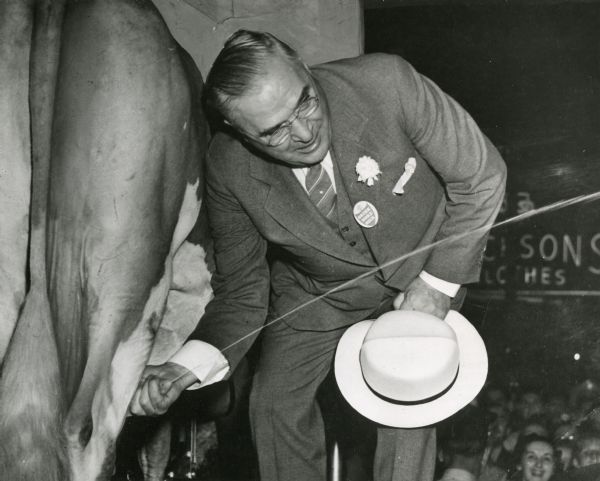 Governor Julius Heil squirting milk from a cow on a parade float. The Governor climbed aboard the float during the dairyland parade, a part of Milwaukee's celebration of Wisconsin Products Week. He is wearing a flower and a button reading "drink more milk" on his lapel. A portion of the crowd can be seen at bottom right.
In the background is Jackson's Clothes at 180 W. Wisconsin Avenue.