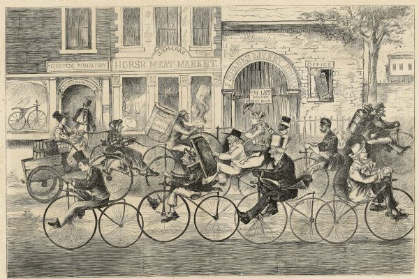 Humorous engraved image showing a street scene crowded with people riding bicycles. In the background are businesses identified as J. Shanks Livery Stable, which appears to be in disrepair and which has a "to let" sign on the door. Next door is J. Shanks Horse Meat Market, and to the left of that is a velocipede manufactory and riding school. A man is exiting the riding school with his arm in a sling.