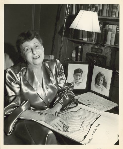 Irna Phillips is seated, and drawing a floor plan. On a table next to her are framed photographs of her children.