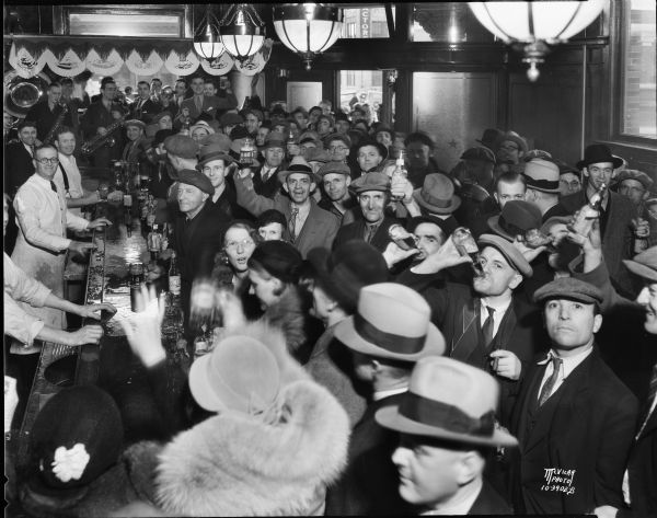 Slightly elevated view of crowd in the Fauerbach Brewery tavern at 651 Williamson Street, with men and women drinking and toasting and celebrating the end of Prohibition. A small band plays in the background.