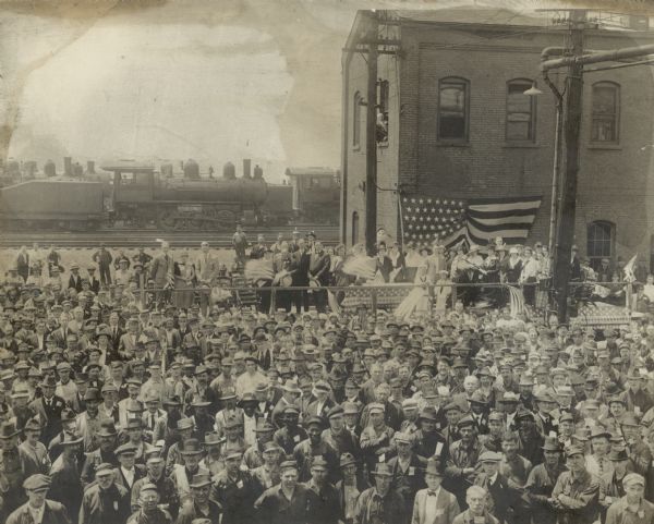 Elevated view of crowd at flag raising ceremony at Western Avenue. A group of people are standing on a stage or platform decorated with flags, and a large crowd, many with miniature flags, gathered in the foreground. In the background is a brick building and locomotives on railroad tracks.