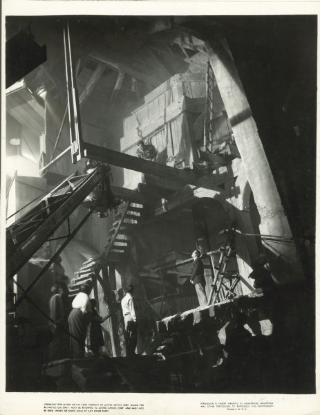 A behind the scenes photograph of the set of the film "Foreign Correspondent." Joel McCrea is on a platform high above a group standing below. Alfred Hitchcock is standing at the bottom of the stairs.