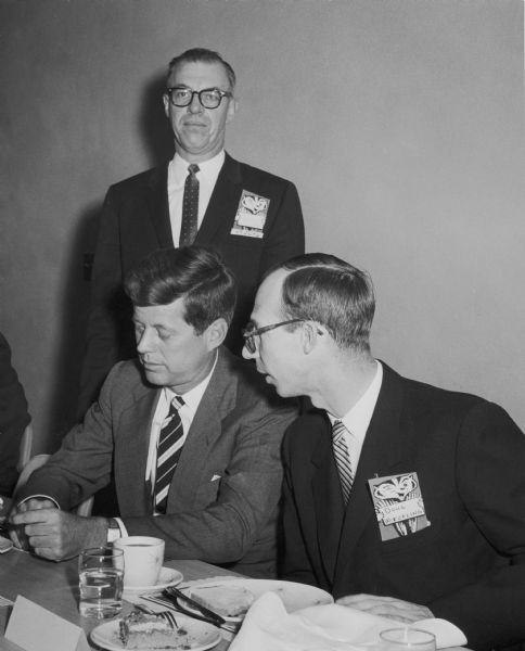 John F Kennedy John F. Kennedy seated next to Doug Witzeling at an event for the Chamber of Commerce. A man stands behind them.