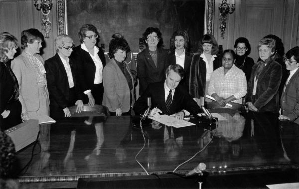 Governor Tony Earl at the signing of the executive order creating the Wisconsin Women’s Council. At the end of the remarks he invites women from the legislature and his administration to join him for the signing. 
Pictured from left to right:
1. Sen. Susan Engeleiter
2. [not identified]
3. Eileen Mershart (Deputy Secretary DOR)
4. Doris Hanson (DOA Secretary)
5. Rep. Barb Gronemus
6. Rep. Mary Lou Munts
7. Rep. Sharon Metz
8. Rep. Barb Ulichny
9. Rep. Marcia P. Coggs
10. Linda Reivitz (DHSS Secretary)
11. Rep. Lolita Schneiders
12. Roberta Gassman (Policy Advisor Governor’s Office)
