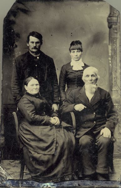 Studio portrait of family of four in front of a painted backdrop. An older man and a woman sit in front, and a younger man and woman stand behind them.