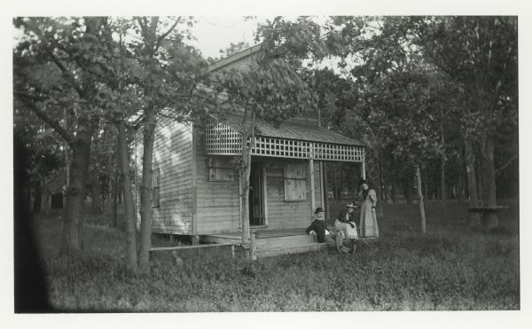 Exterior view of Oak Lodge near Lake Mendota, with a man and two women posing on the front porch. It was owned by Caroline Fitch, Ann Stewart, and Jessie Vilas.