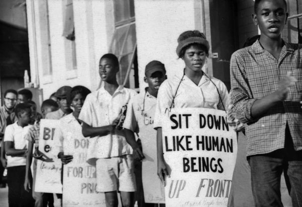 Demonstration at the Greyhound Bus station, showing a woman holding a sign that reads: "Sit Down Like Human Beings, Up Front." SNCC Arkansas Project.