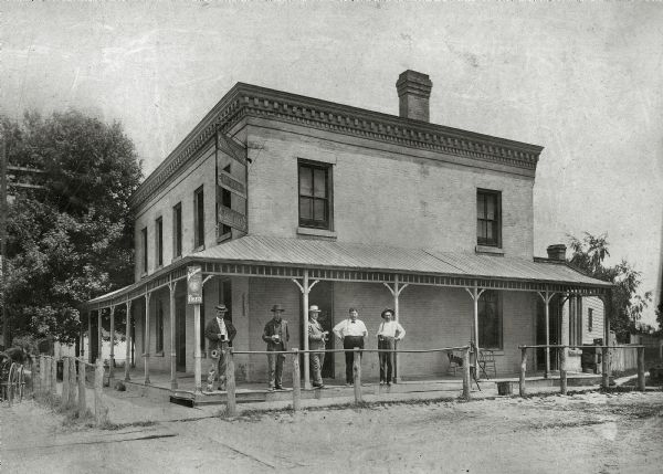 The Union House Tavern, known as Schulkamp's Corner Saloon, 2601 East Washington Avenue at Milwaukee Street. A group of men are standing on the porch holding glasses of beer. There is a hand-pump and a pail on the wooden sidewalk running in front of the porch. There is a horse and wagon tied up at the hitching post on the left. A sign for Breckheimer Beer is on the corner of the building above the porch roof.
