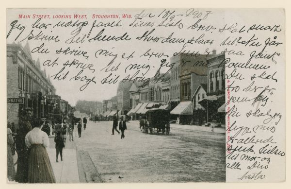 View along sidewalk of Main Street, with pedestrians on the sidewalk and walking across the street. Storefronts line both sides of the street. Caption reads: "Main Street, Looking West, Stoughton, Wis."
