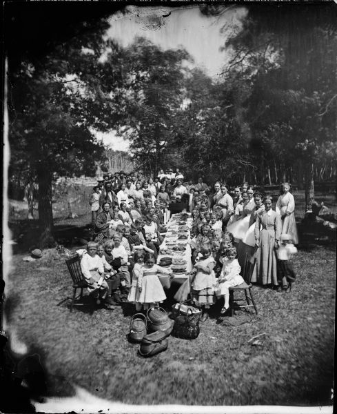 Slightly elevated view of a large, multi-generational Norwegian family and friends gathered for a picnic celebration. In the foreground are picnic baskets on the ground at the end of a long table laden with plates of food. Children holding diplomas are seated in chairs along the side of the table. Adults are posed around the table. A woman holding a bell sits behind another table stacked with books in the center of the group. Women and men stand around the sides.