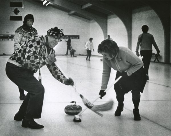 L.J. Brodd and Mrs. Robert Rasche, both of Glendale, sweep ahead of the stone during a match as part of the 21st Annual Milwaukee Mixed Bonspiel. Mrs. Jane Brodd is watching in the background.