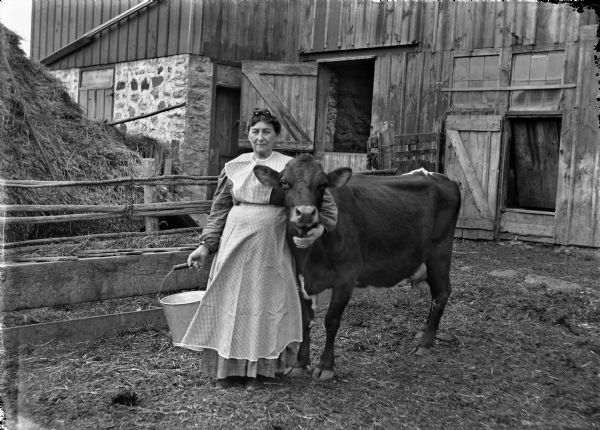 Grandma Martha (Goetsch) Buelke posed in front of a barn with her favorite cow. She is holding a milking pail.