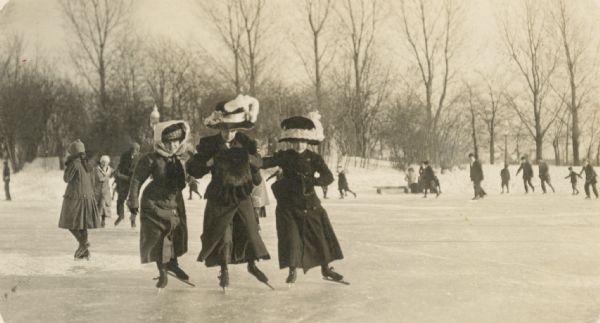 Three women wearing large hats ice skating together on a pond.