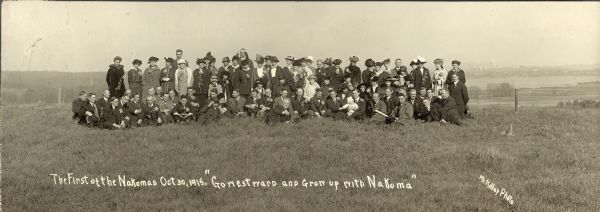 Outdoor group portrait of people identified as "settlers" (lot buyers) to the Nakoma development. Many of the people are wearing or holding a long-stemmed flower. The Wisconsin State Capitol is in the far background across Lake Monona. At the bottom is written: Go westward and grow up with Nakoma. 