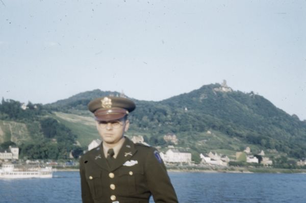 John Scott in Uniform Outdoor portrait of a uniformed man, probably John Scott. Behind him is a boat on a body of water, and on the opposite shorelineis a town and hillside. Taken during Armed Forces trip in Europe and North Africa.