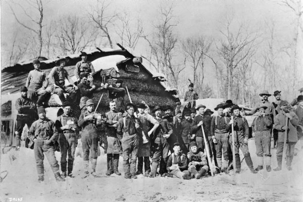 Group portrait of a large logging crew posing outdoors in the snow at their camp in front of a log building. One of the men is playing a fiddle or a violin, and two men are posing with their fists up as if ready to fight. One of the men sitting on the ground in front of the group appears to be holding a dog.