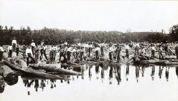 Log driving crew for the Chippewa Lumber & Boom Company. Crew members are in the process of breaking a jam at the Big Eddy. One of the men is carrying a young boy on his shoulder.