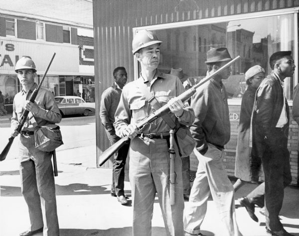 Civil rights activities near the Canton, Mississippi courthouse. Two police officers are pictured holding shot guns and carrying bags of tear gas equipment. The African-American man wearing a hat and with his face partially obscured by the barrel of a gun is identified as Joe Lee Watts, CORE (Congress of Racial Equality) task force worker.