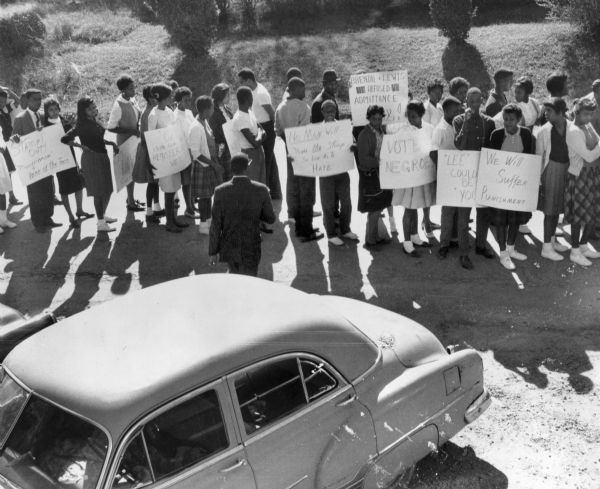 Elevated view of a group of students from McComb High School standing near a car holding signs with messages, including: "Vote Negroes" and "We Will Suffer Punishment."