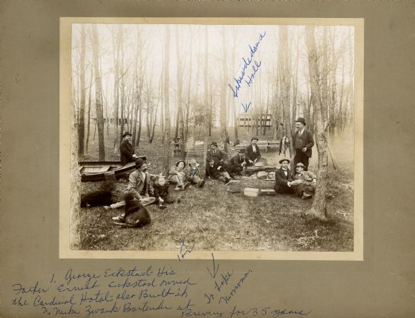 Fishing Outing A group of men relax outdoors after fishing. Most are seated on the grass, while one man sits on and boat and another stands holding the day's catch. There are fishing poles propped up against trees behind them. The man on the left is sitting with two dogs and a child. The third man from the left is George Eckstad. The fourth man from the left is Mike Zwank (Swank), who was the bartender at Fauerbach Brewing Company for 35 years. Several other men pictured here are probably from the Fauerbach family. Karl Fauerbach later wrote captions on this photograph in blue ink.
