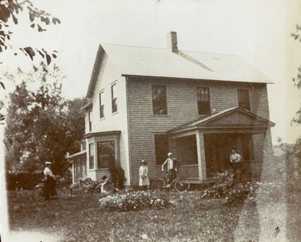 John Lloyd Jones house, near Hillside Home School on Highway 23 across from Taliesin. A man and a woman are standing and holding bicycles, one woman is kneeling by flowers, one woman is standing against the house and another man is standing on the porch.
