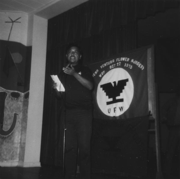 Cesar Chavez speaking while standing in front of a United Farm Workers banner commemorating the unionization of workers at a flower nursery on October 22, 1975.