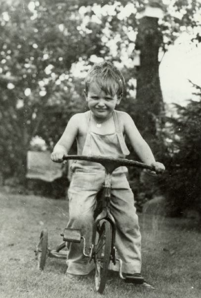 Boy Riding a Tricycle A boy wearing overalls is riding a tricycle outdoors. A caption identifies him as John Peters.