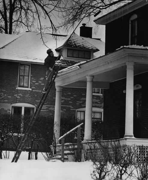 A man on a ladder is using a snow rake to rake his roof. The ladder is propped against the portico of his house. Caption reads: "PROBLEM SOLVED — Homeowners have watched and worried about the buildup of snow on roofs this winter, but Marvin Franz of Chippewa Falls acted. He simply raked it off."