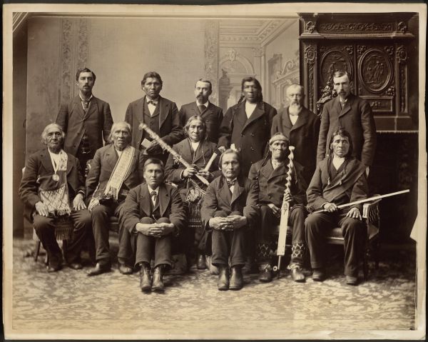 Group portrait of Ojibwa Chiefs from Bad River, Lac Courte Oreilles, and Lac du Flambeau, in Washington, D.C. The reason they were there is in dispute. Records show it had something to do with a bill to divide the reservations into parcels, reducing the size of the reservations and opening the lands for logging. Names, left to right, seated on floor: Wadwaiasoug, Oshawashkogijig. Seated in chairs: Edawigijig "Both Sides of the Sky" (Bad River Chief and signer of 1854 Treaty), Kiskitawag or Giishkitawag "Cut Ear" (signed multiple treaties as a warrior of the Octonagon Band, but afterwards was associated with the Bad River Band), Akewainzee (most prominent Chief of the Lac Courte Oreilles band), Nizhogiizhig "Second Day," Oshoga. Standing: Vincent Conyer (interpreter), Ogimagijig, Dr. I.L. Mahan (Indian Agent in Bayfield, 1878-1879), Wasigwanabi, George P. Warren (Civil War veteran, Private, Company K, 36th Infantry, he was born on 1824 in LaPointe, wounded at Cold Harbor on 6-3-1864, mustered on 3-11-1865), and Thaddeus Adams Thayer, Jr. (married to Mary (Bert) Thayer "Gagi," an Ojibwa woman). They are posing in front of a painted backdrop.