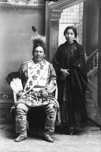 Studio portrait in front of a painted backdrop of Alex Lonetree, sitting and his wife, standing. They came to live with the Winnebagos after the last removal to Nebraska.