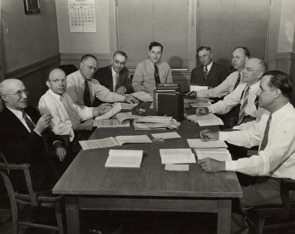 Nine men are sitting around a table in an office, with books and papers in front of them. A caption identifies them as, from left to right: August Alles, Paul Grummel, Al Hoffmann, John Klatt, Henry Lengling, Art Olsen, Cy Mott, Otto Jirikowic, Ed. Madsen.