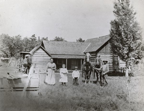 A man, his wife and their two children are posing in front of their farmstead. The man is holding a team of horses. Another horse pulling a buggy is in the background on the left.