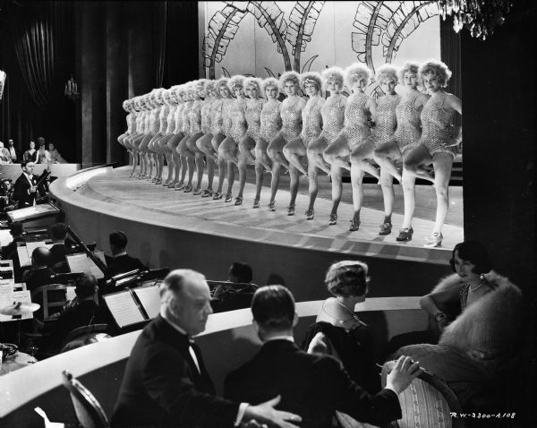 Chorus Girls in the Film "Alibi" Chorus girls standing on stage in a scene from the 1929 film "Alibi." Each dancer is standing on one leg with the other leg bent at the knee. The orchestra is in a pit in front of the stage. Two men and two women are sitting in a box seat near the stage.