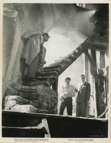 Scene Still from "Foreign Correspondent" Scene still from the 1940 film "Foreign Correspondent." Mr. Krug (Eduardo Ciannelli) stands with his hand on the shoulder of Tramp (Martin Kosleck) underneath a staircase inside a windmill. John Jones (Joel McCrea) stands on the steps of the stairs to the left of the two and tries to stay out of sight.