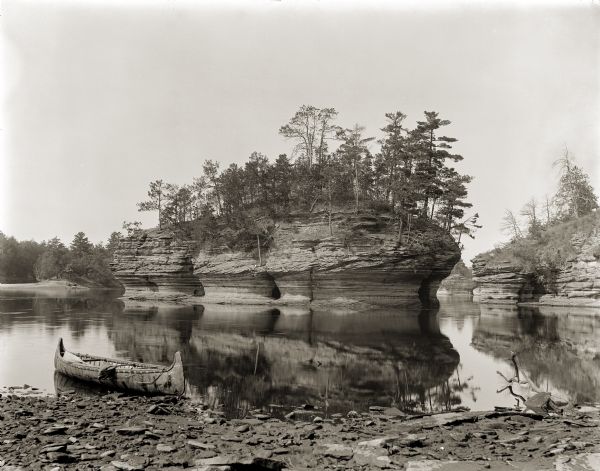 Lone rock viewed from the shore; there is an empty canoe drawn up on the shore in the left foreground.