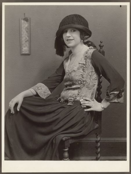Helen Ferguson sitting in chair and smiling Actress Helen Ferguson sits in a high back chair and smiled towards the camera. She has one hand on her hip and the other resting on her knee. She wears a hat with fringe on one side and crepe and satin dress with buckles along with a single strand pearl necklace.