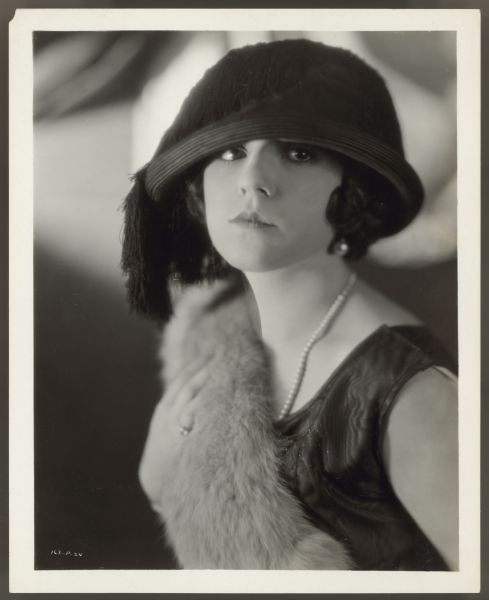 Helen Ferguson in black hat Actress Helen Ferguson looks directly at the camera in a photograph by Clarence Bull. She wears a black hat that covers her forehead and has fringe on the side. One earring can be seen along with a single strand pearl necklace. Her dress is sleeveless and she has a fur stole draped over one shoulder.