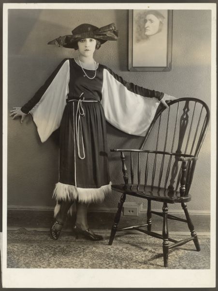 Helen Ferguson in a black dress with monkey fur Actress Helen Ferguson poses against a wall win a black dress with white monkey fur. Her hat has a very large bow on the front. One hand rests on the top of a Windsor chair. A framed picture of a man's head is on the wall behind her.