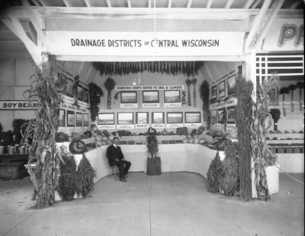 Drainage Districts of Central Wisconsin booth at Wisconsin State Fair with crops on display, and photographs around the booth above vegetables on display on tables. There is a man sitting in the booth.