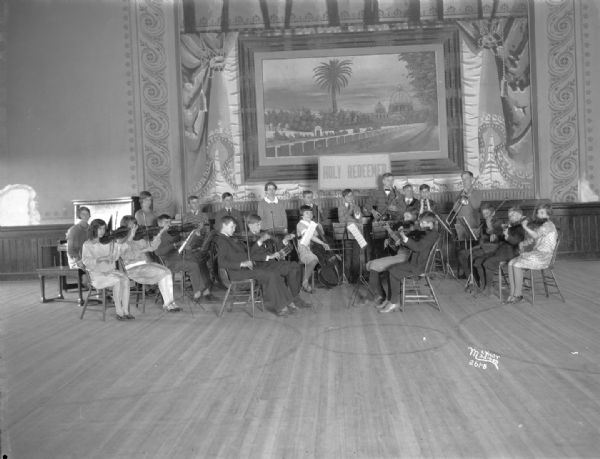 Group portrait of the Holy Redeemer school orchestra, 128 W. Johnson Street. On the wall in the background is a large mural surrounding a painting.