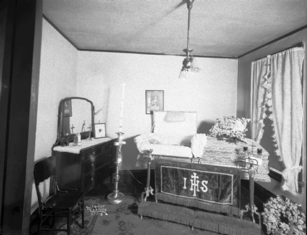 View towards a body in a coffin in a home with a "Mother" floral arrangement. A cloth in front of a religious kneeler in front of the coffin reads: "IHS."