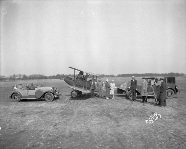 Mr. Sutton and Fay directing a scene for the movie "American Girl." Eight people are standing in front of an Oakland automobile and airplane, and four more people are sitting in another Oakland automobile at Pennco Field (Royal Airport). The tail of plane reads: "Royal Airways Corp."