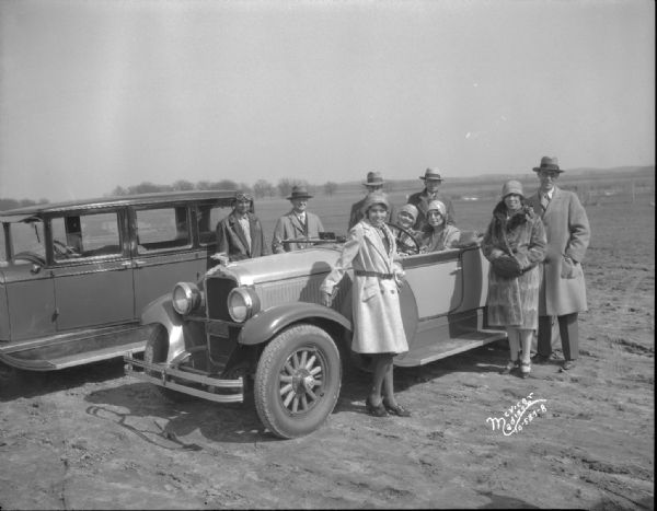 Group of four women and four men and an airplane pilot standing in and around two Oakland automobiles at the Pennco Field (Royal Airport). Taken for Associated Motors Inc.